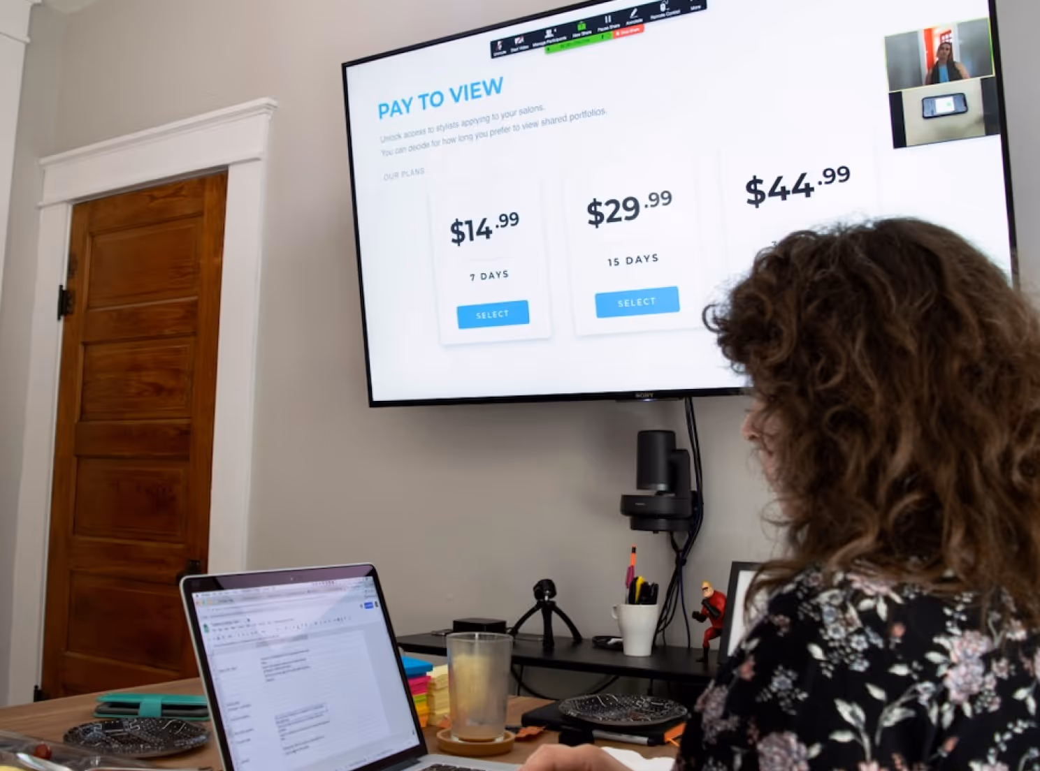 Woman working on laptop in front of a wall screen showing subscription plans with prices and video call window.