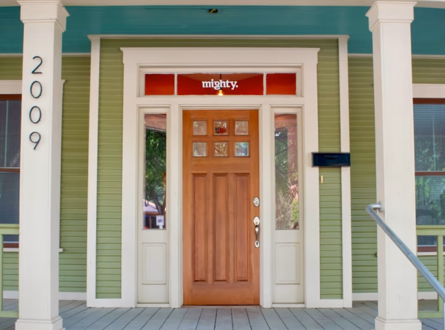 Front porch with green siding, wooden door, white trim, house number 2009, and 'mighty.' sign above door.