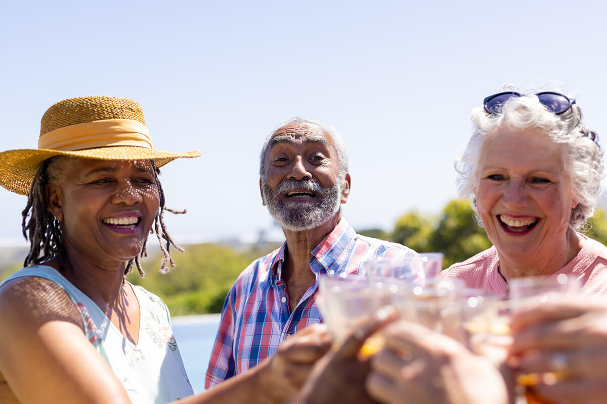 Three senior adults smile and raise glasses in a toast on a bright, sunny day.