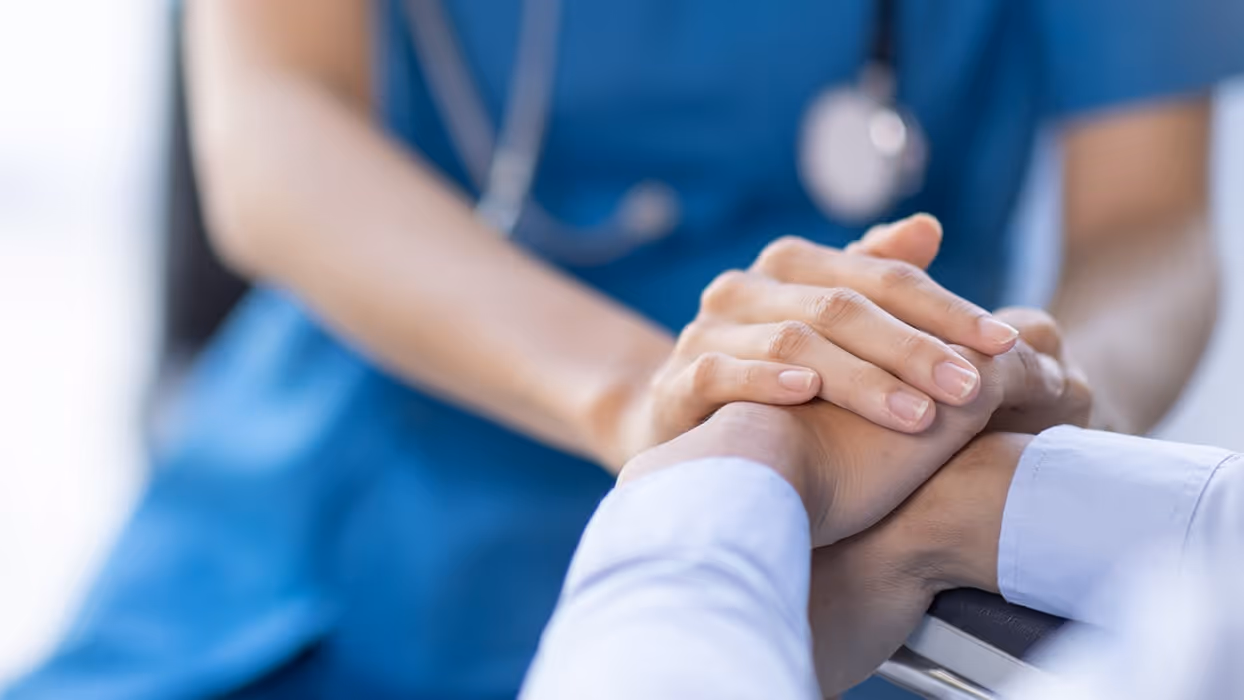 the hands of a blue-scrubs-clad doctor, with a stethoscope visible around their neck, holds those of a patient. We can only see the hands and arms of each, and the body of the doctor.
