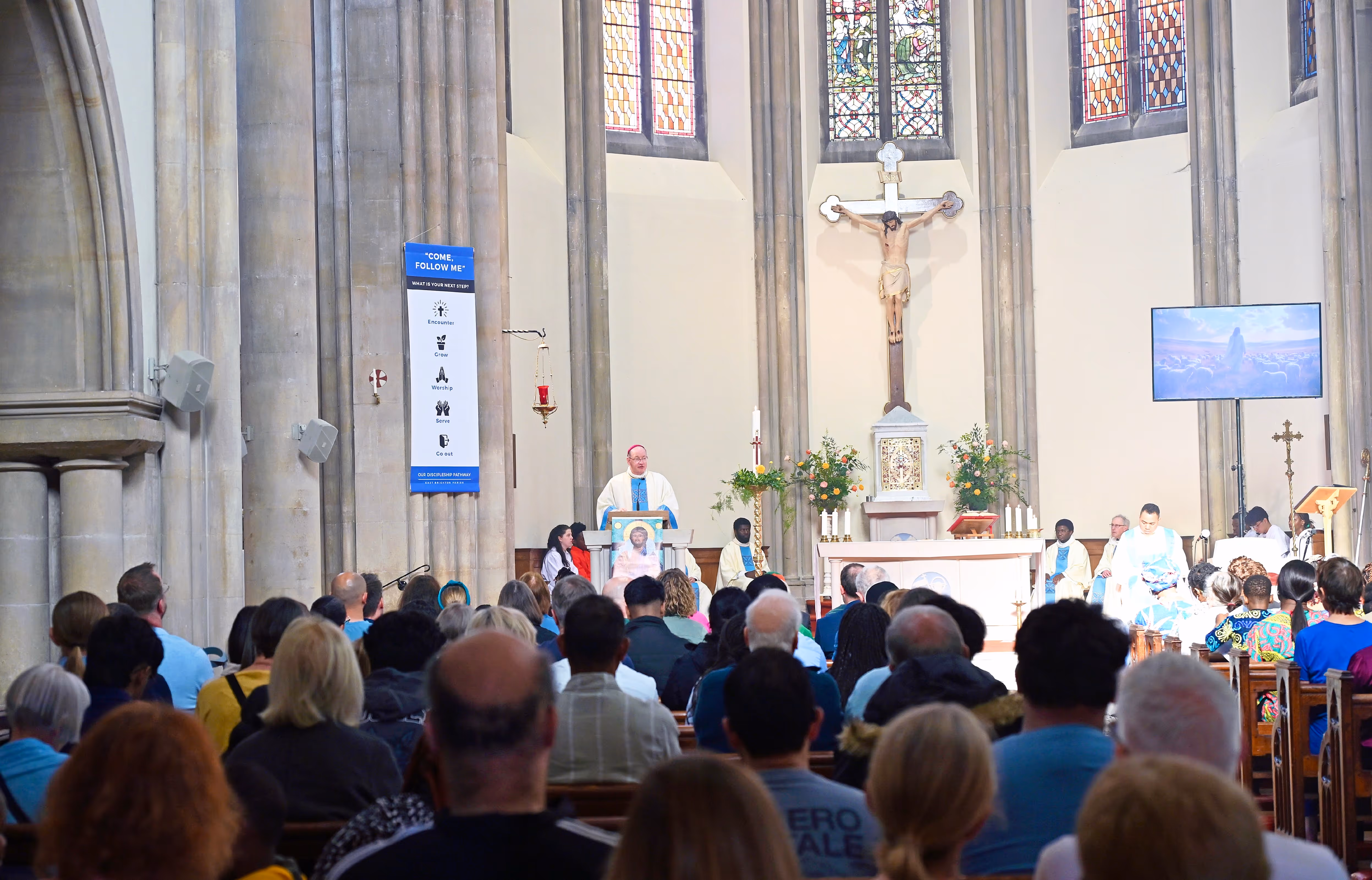 A view over the heads of a large congregation in St Joseph's, Brighton, a stone-built church with white-rendered walls, towards Bishop Richard who is at the ambo. Other clergy can be seen seated around the sanctuary. A very large crucifix is situated above the tabernacle, behind the altar.