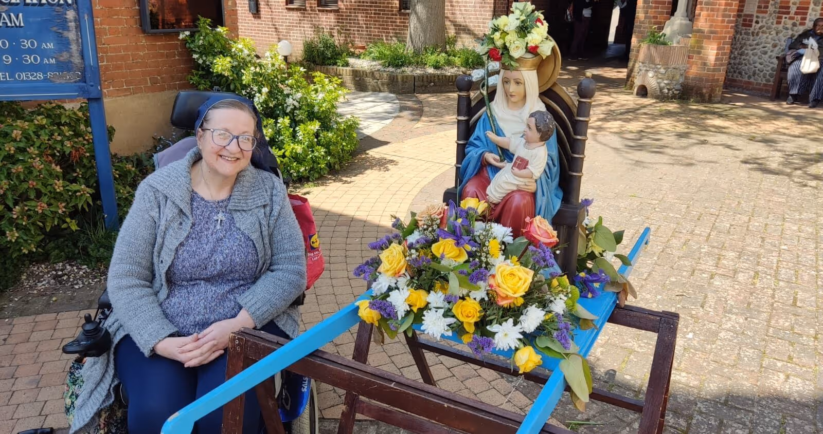 Sister Marika sits in a courtyard, in a powered wheelchair. She wears a veil. Beside her is a statue of Our Lady on a carrying platform and adorned with flowers.