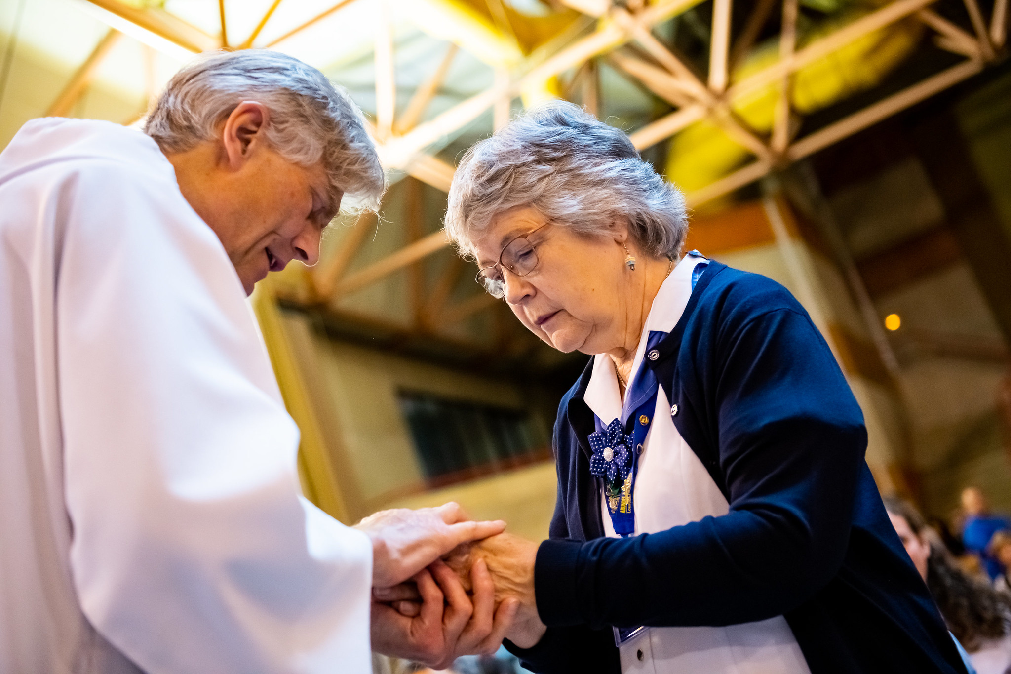 Canon Tom Treherne, eyes closed, blesses the hands of a helper in uniform, holding her hands between his.