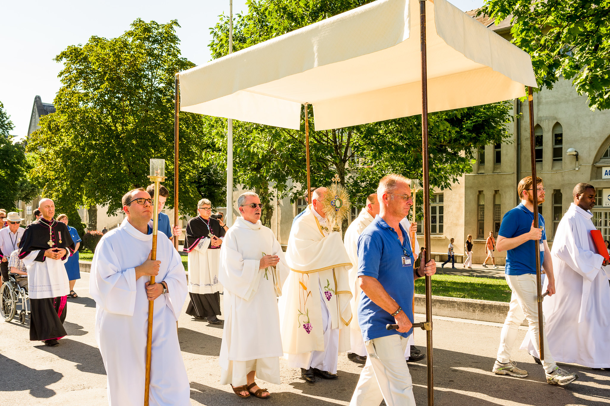A monstrance holding the Blessed Sacrament is carried by Bishop Richard on the esplanade under a canopy borne by four helpers, and accompanied by servers with lighted candles. Other clergy follow.
