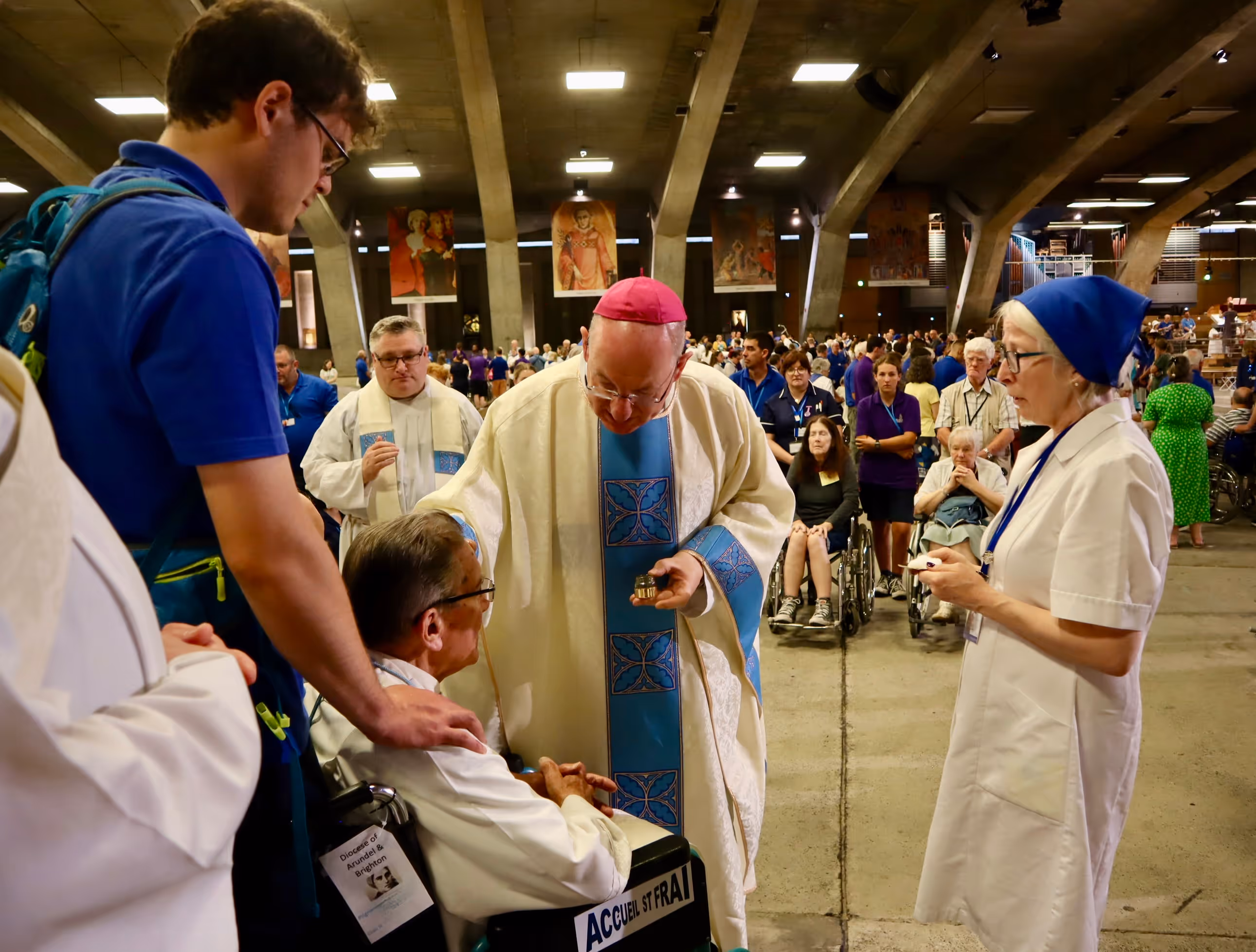 Bishop Richard is pictured in the Underground Basilica, bending over Canon Seamus Hester, whose forehead he is anointing with oil. Mary C stands by, holding cotton wool for cleaning his fingers, and a helper holds Canon Seamus’ wheelchair. Fr Simon Dray is looking on from a distance and other pilgrims can be seen in the background.