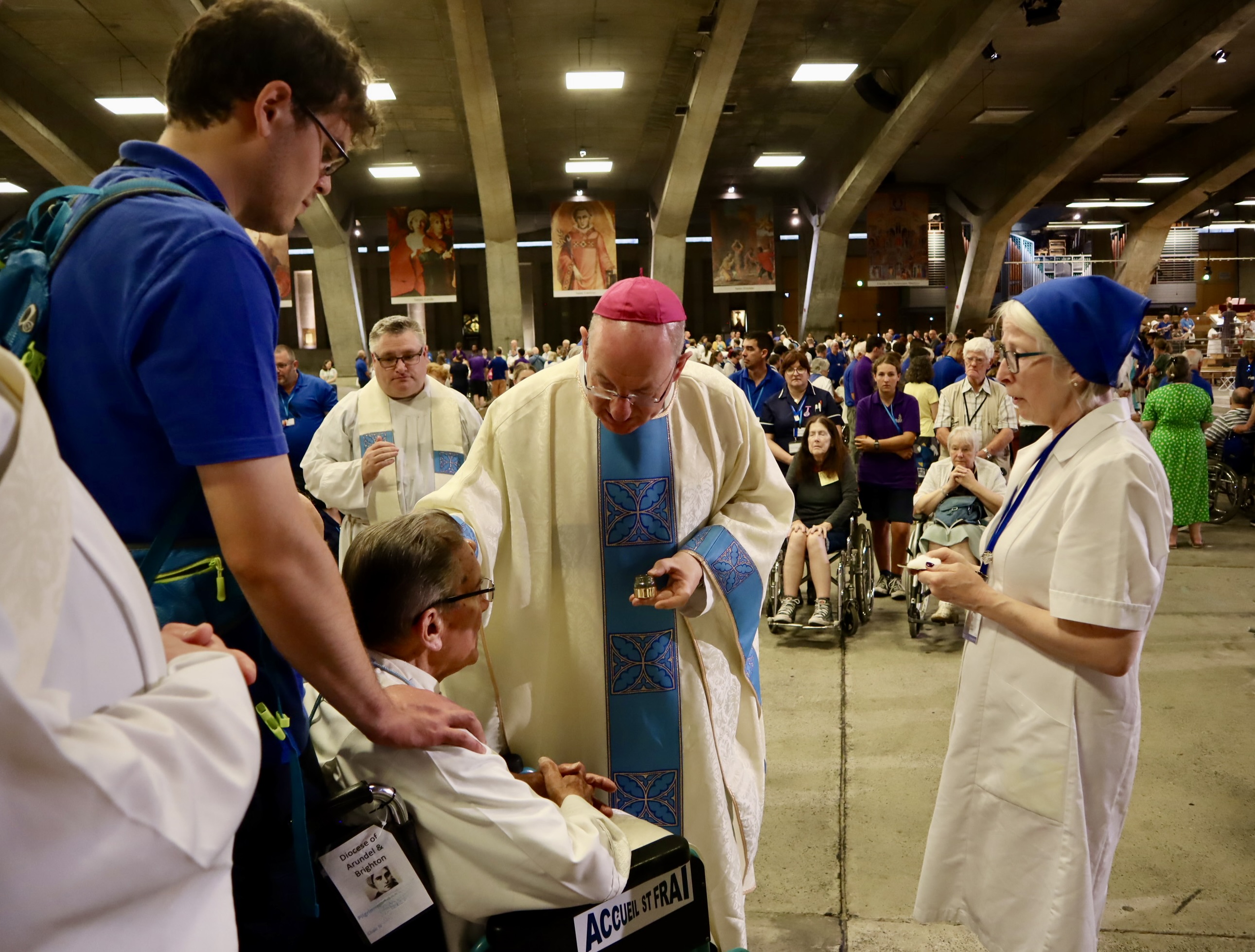 Bishop Richard is pictured in the Underground Basilica, bending over Canon Seamus Hester, whose forehead he is anointing with oil. Mary C stands by, holding cotton wool for cleaning his fingers, and a helper holds Canon Seamus’ wheelchair. Fr Simon Dray is looking on from a distance and other pilgrims can be seen in the background.