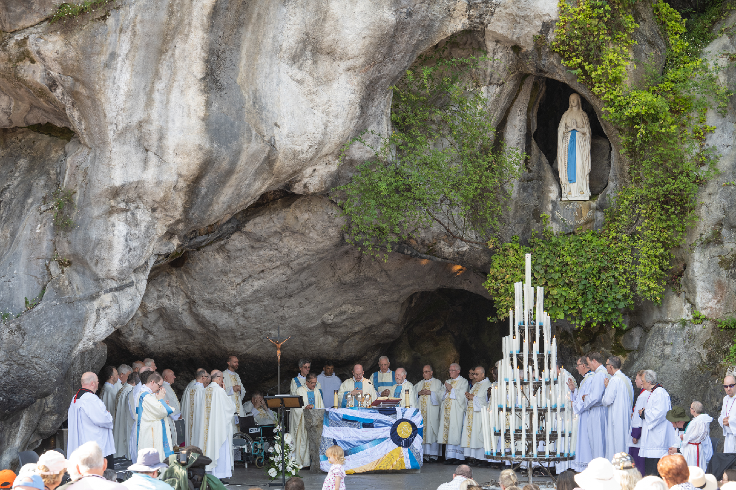 Vested in white, Bishop Richard and the priests and deacons in Lourdes are pictured in the Grotto, beneath the statue of Our Lady. A colourful altar cloth covers the altar.