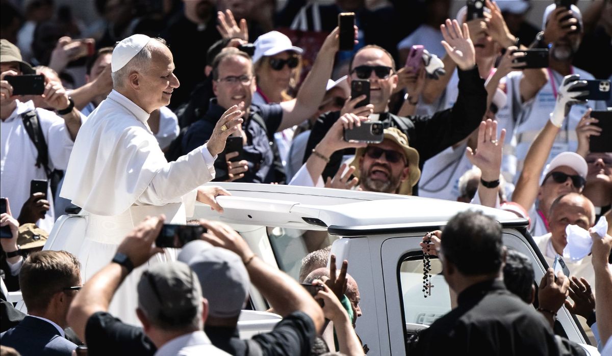Pope Leo is pictured standing in the back of a popemobile, smiling and waving, surrounded by people holding up phones and cameras.