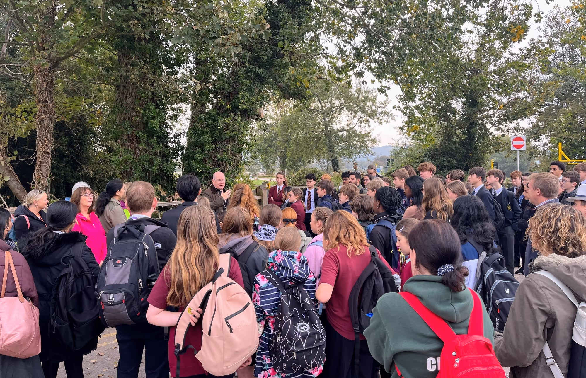 Dozens of students wearing coats and backpacks are pictured gathered around Bishop Richard in a wooded area. He is speaking to them animatedly, gesturing.