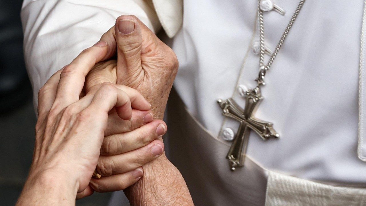 Photo shows the hand of a man in a white cassock, wearing a pectoral cross, grasping tightly another pair of hands that look careworn and elderly