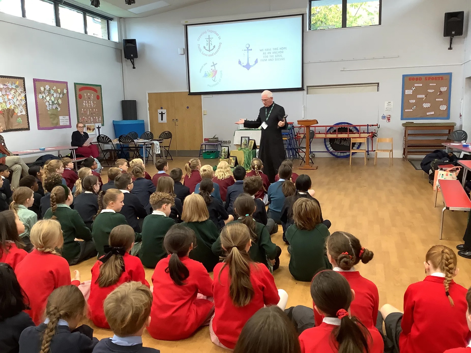 Photo shows Fr Chris Bergin in a black cassock, wearing a lanyard, speaking to a school hall full of pupils wearing various different uniforms. Visible on the whiteboard behind him are the jubilee and other logos.