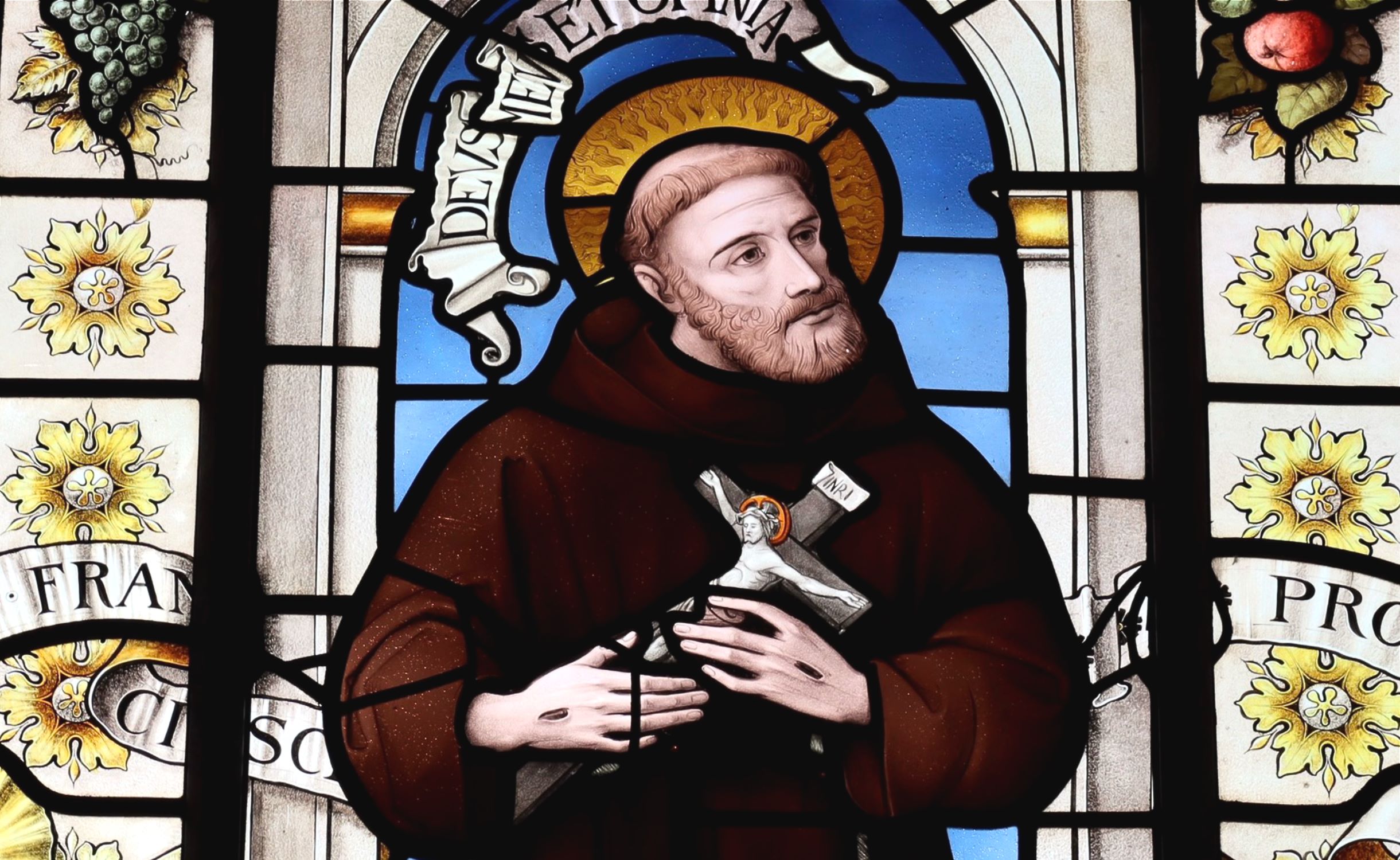 Stained glass window depicting St Francis in a brown habit, with a halo, holding a crucfix, and looking away to our right.