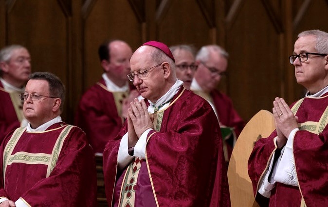 Archbishop Richard is pictured in prayer, vested in red and flanked by two deacons.