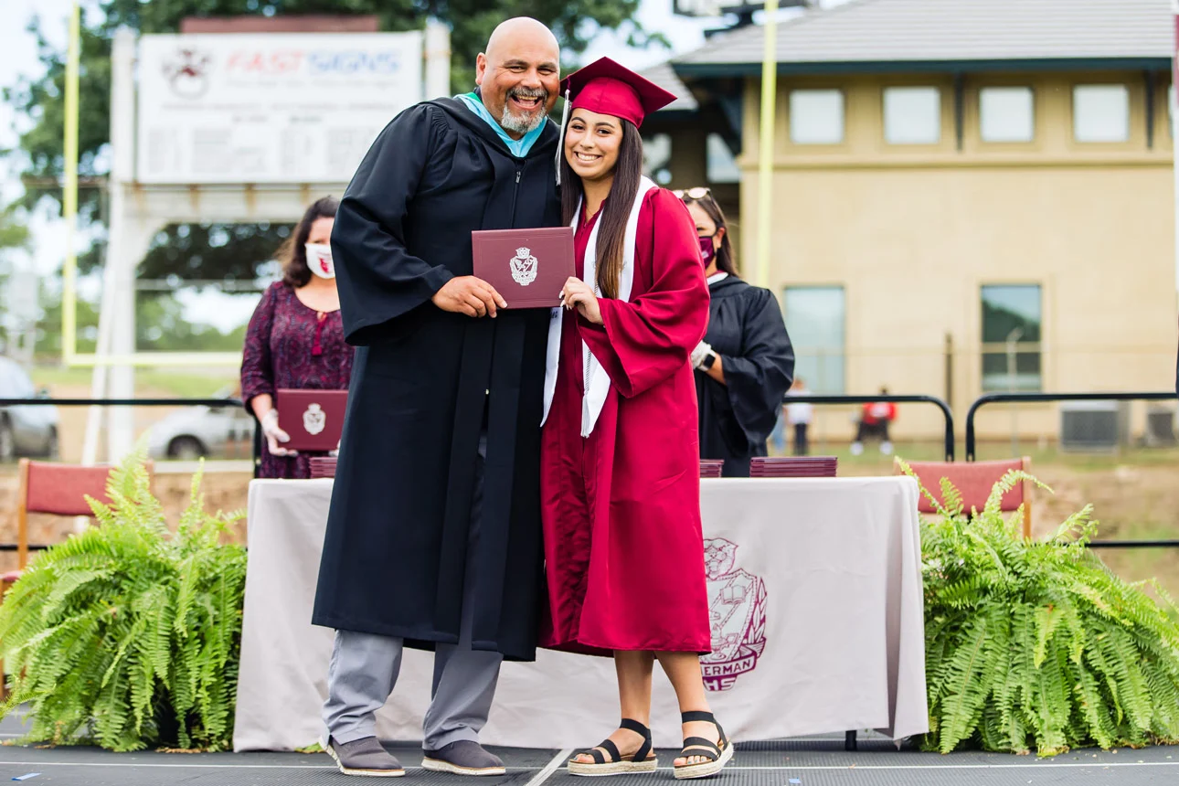 Graduate in maroon cap and gown smiling and holding diploma with faculty member in academic robe on outdoor stage.