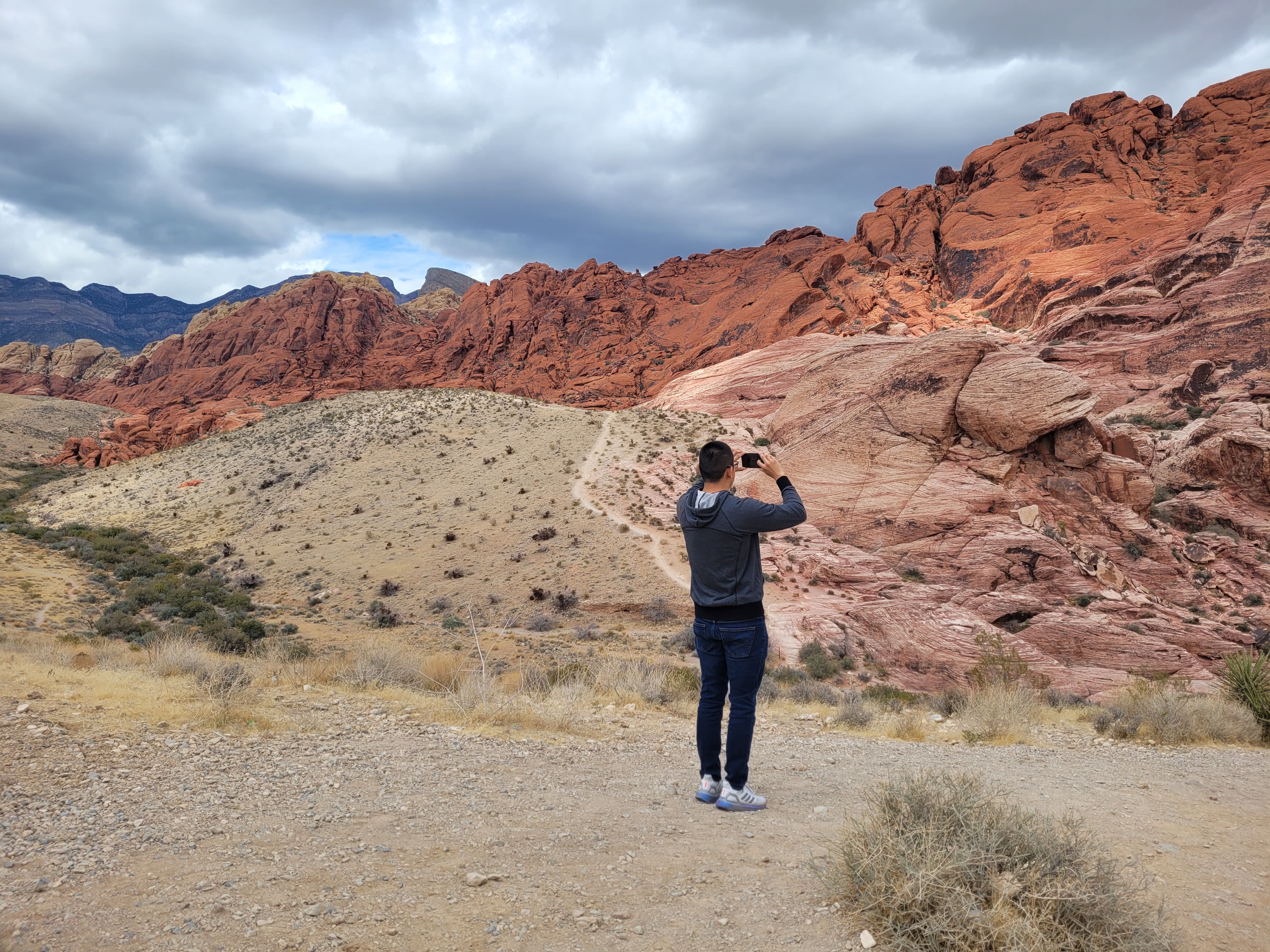 A man is visiting Red Rock Canyon near Las Vegas. He is taking a picture of the swirling red and orange colors of the sandstone canyon. The sky is cloudy and the canyons surround the man.