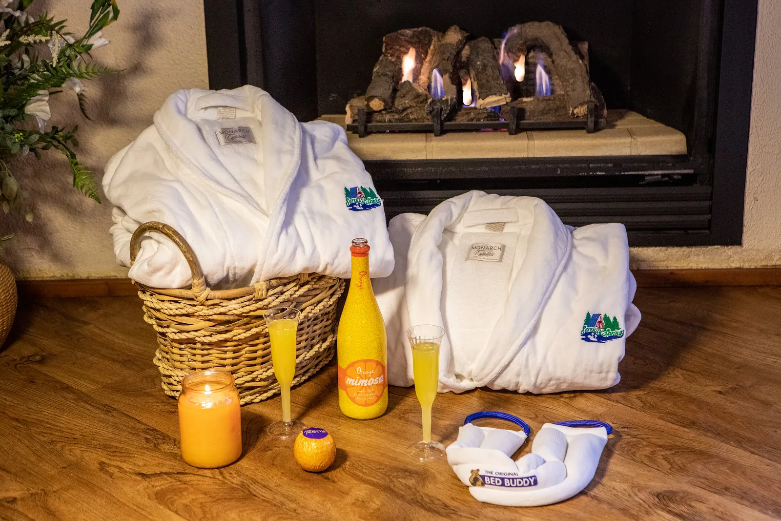 Two white bathrobes with a green and blue embroidered logo placed near a fireplace, accompanied by a bottle and two glasses of orange mimosa, an orange, a lit orange candle, and a white Bed Buddy heating wrap on a wooden floor.