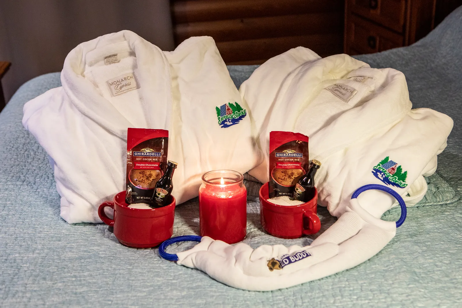 Two white Monarch Express bathrobes on a bed with embroidered Serenity Springs logo, each accompanied by a red mug holding Ghirardelli hot cocoa mix and a small bottle of Baileys, a lit red candle between them, and a white tug toy labeled Tugged Buddy in front.