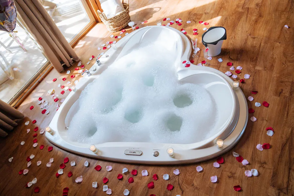 Heart-shaped bathtub filled with bubbles surrounded by red and white rose petals and lit candles on a wooden floor.