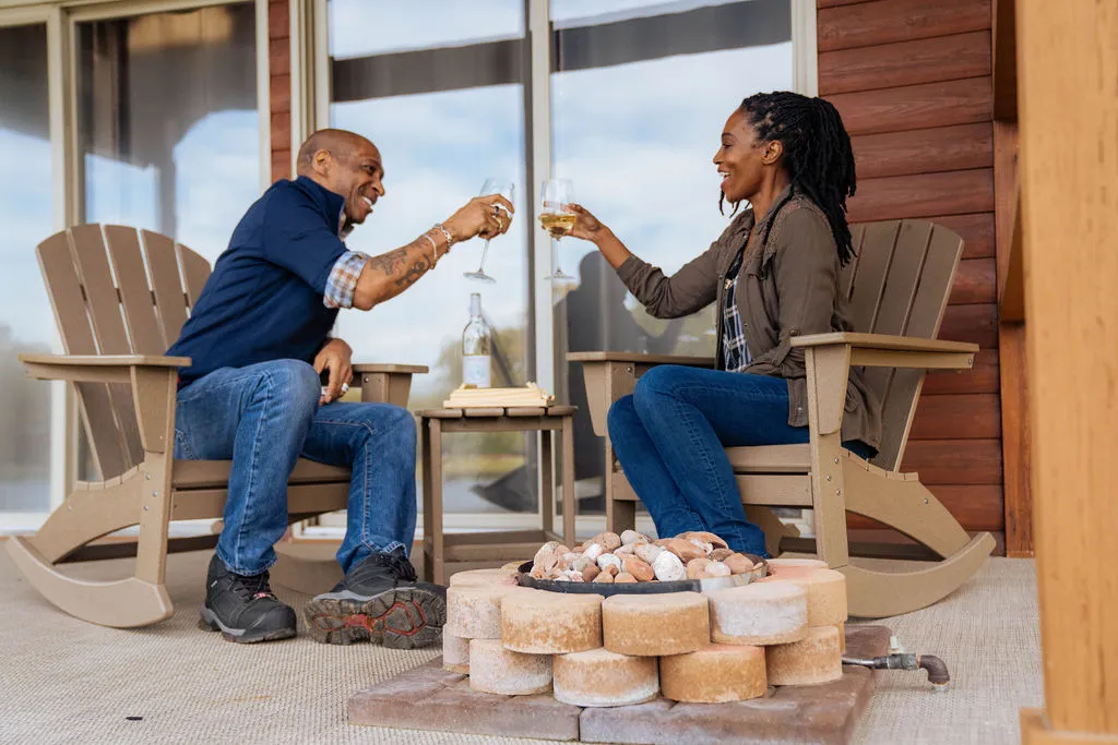 Man and woman sitting on wooden rocking chairs on a porch clinking wine glasses over a small stone fire pit.
