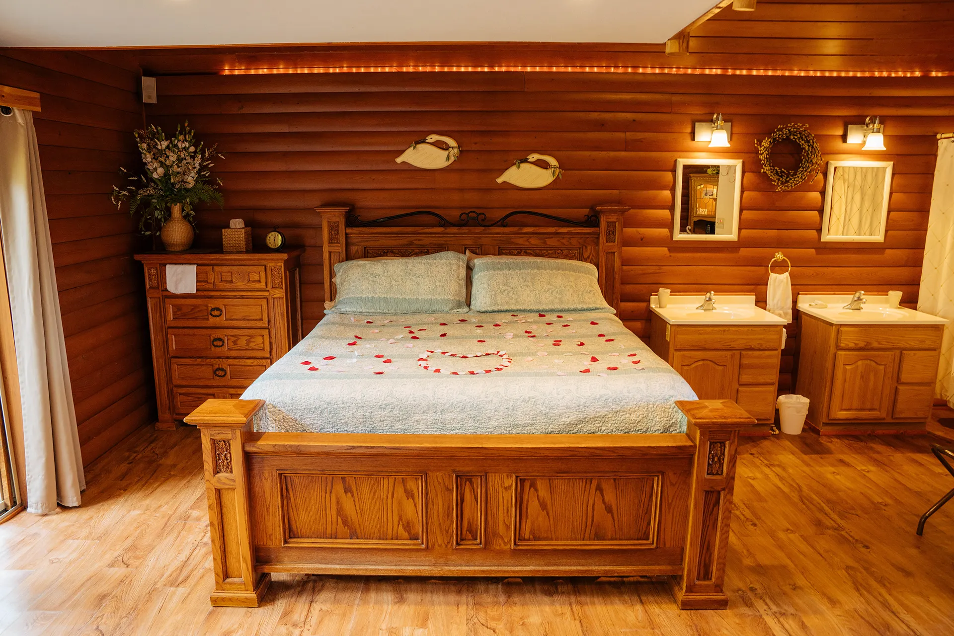 Cozy rustic bedroom with wooden walls and furniture, featuring a large bed decorated with rose petals and two sinks with mirrors on the right side.