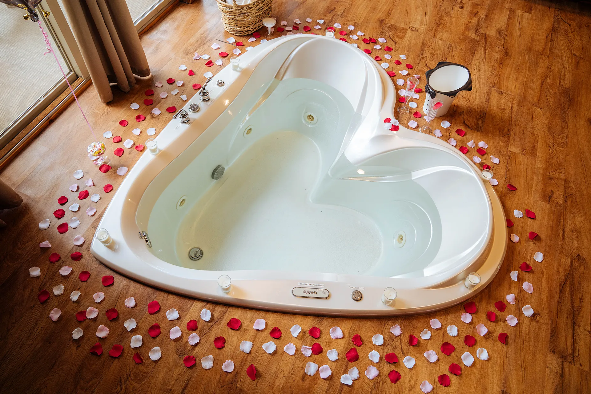 Heart-shaped bathtub filled with water, surrounded by red and white rose petals and small candles on a wooden floor.