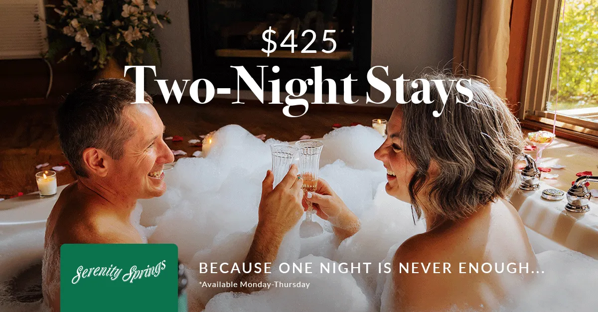 Couple smiling and toasting with champagne glasses in a bubble bath surrounded by lit candles and rose petals.