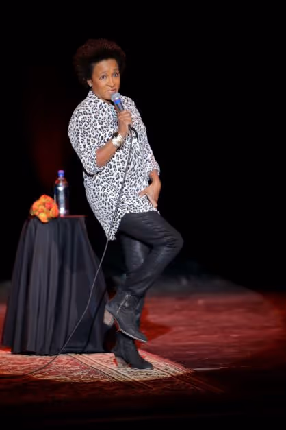 Wanda Sykes illuminated by a spotlight on stage. She has one leg raised as if in the middle of a silly walk. She is speaking into a microphone. 