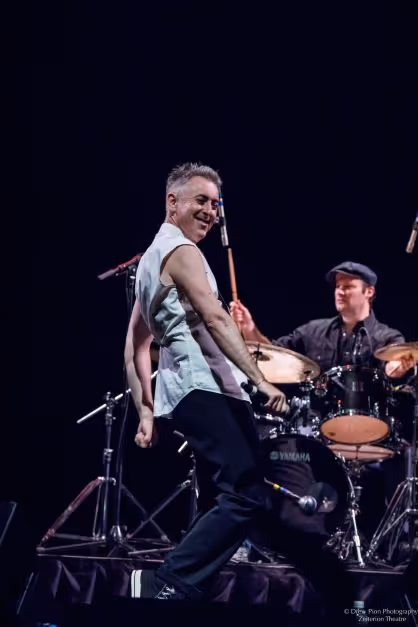 Alan Cummings on stage, mid stride. His body is partially turned away from the camera, and he is looking over his shoulder with a big smile. 