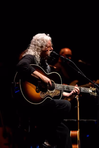 Arlo Guthrie sitting on a stool, playing guitar, and singing into a microphone on a stand. 