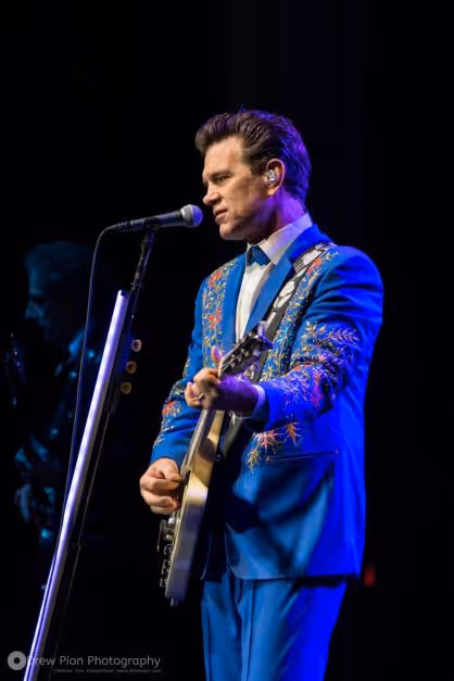 A white man in a blue suit playing guitar and singing into a microphone on a stand. 