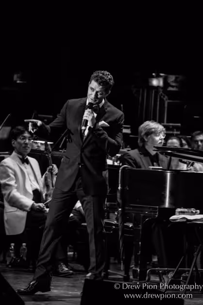 A man standing with a microphone and posed as if he is mid joke with a smile on his face. 