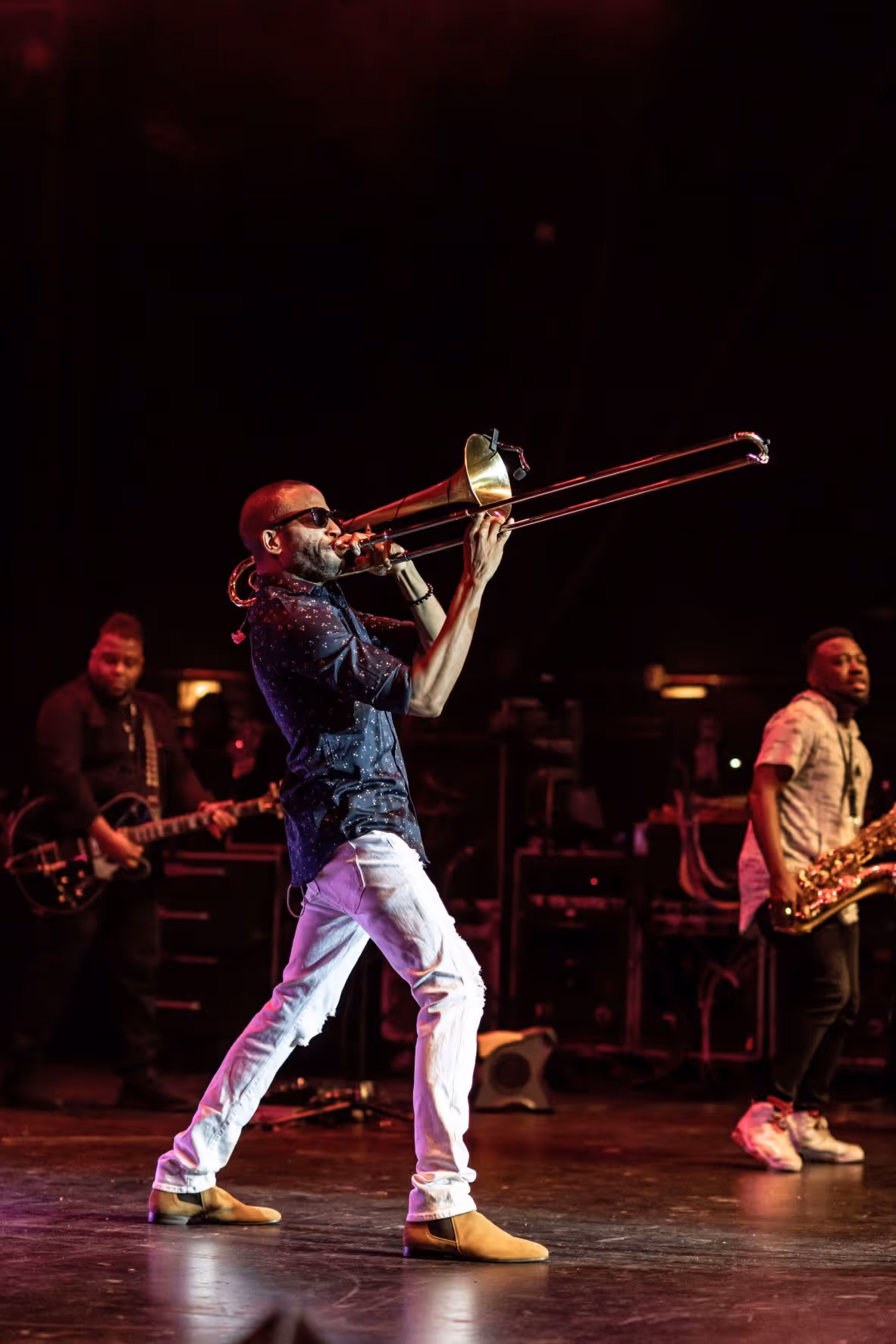 A black man plating trombone on stage. He is wearing sunglasses and is accompanied by a guitarist and bassist.