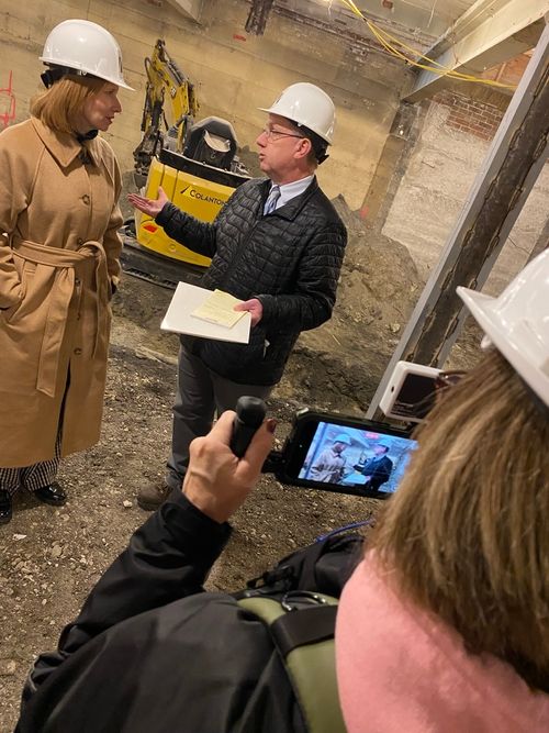 Two people in white hard hats stand on a dirt floor in a construction site, being photographed by a third person.