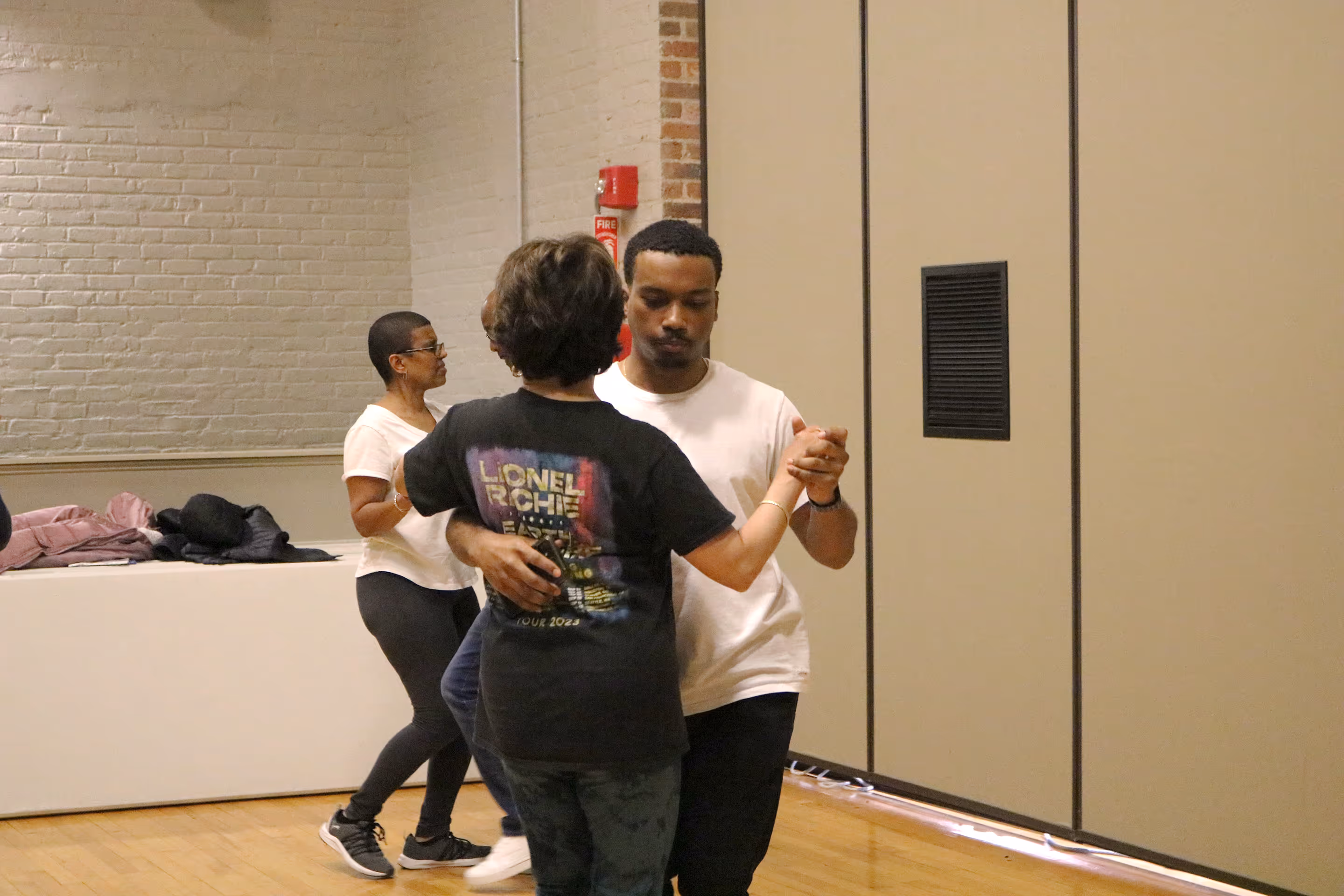 A black man and pale skinned woman dancing together. The woman's back is turned to the camera. Another woman is visible in the background. 