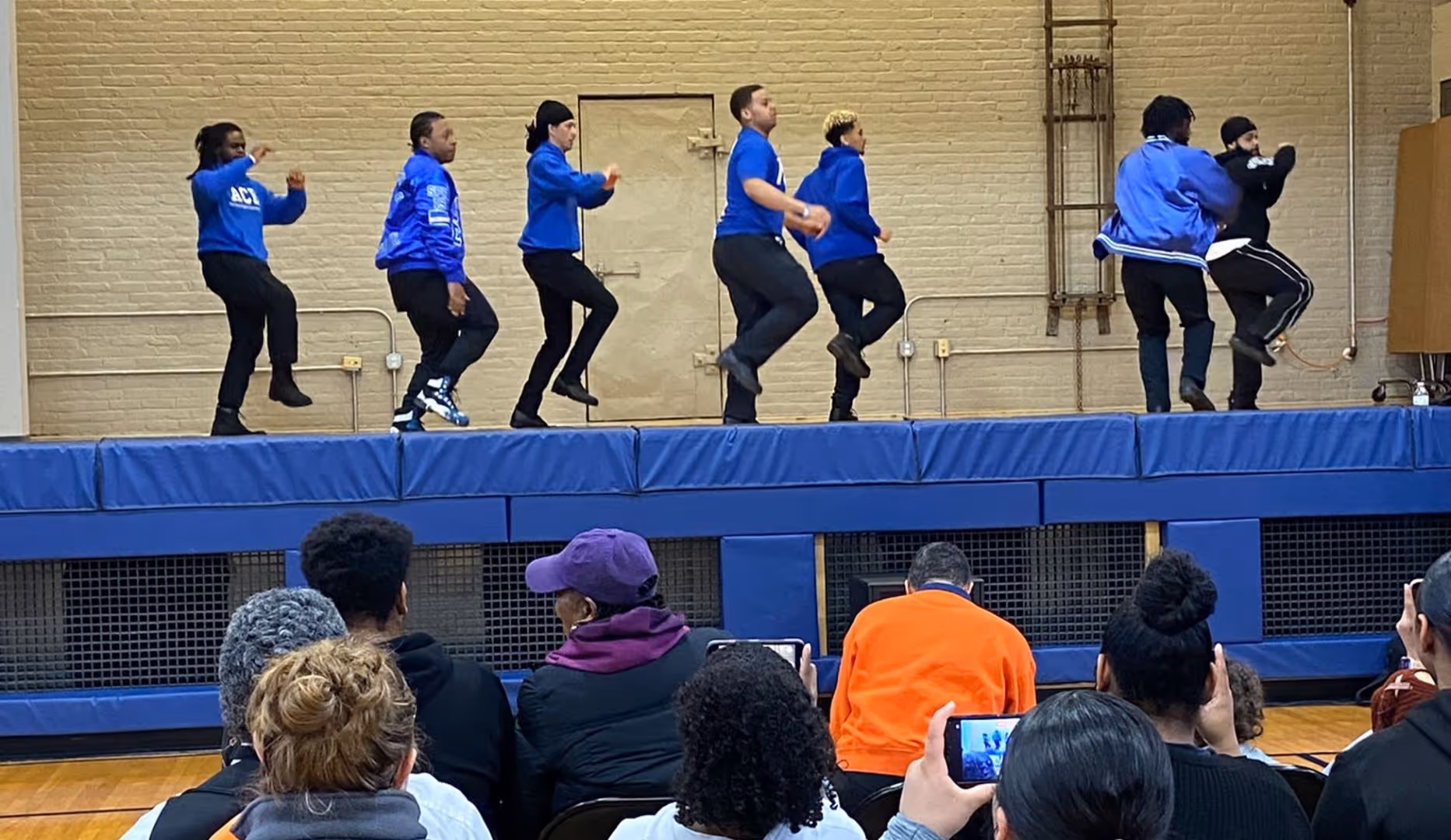 A step dance performance with 7 performers. All are dressed in blue shirts and black pants. 