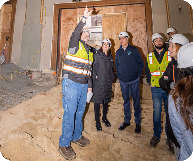 Six people in white hard hats stand in an indoor construction site, atop a sand hill. 