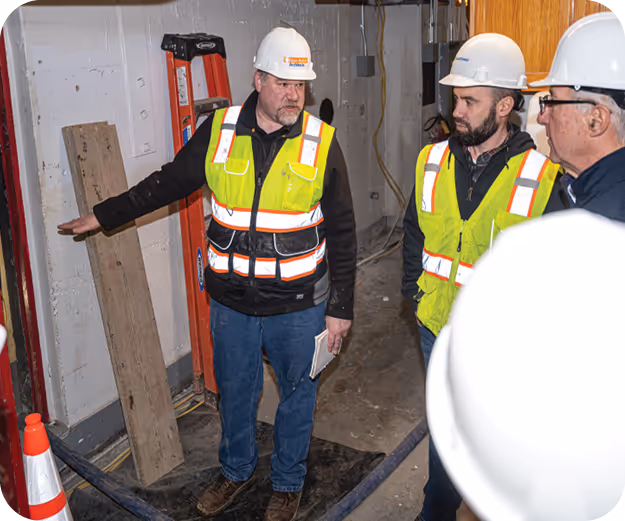 Two men in high visibility vests and white hard hats stand in an indoor construction site. 