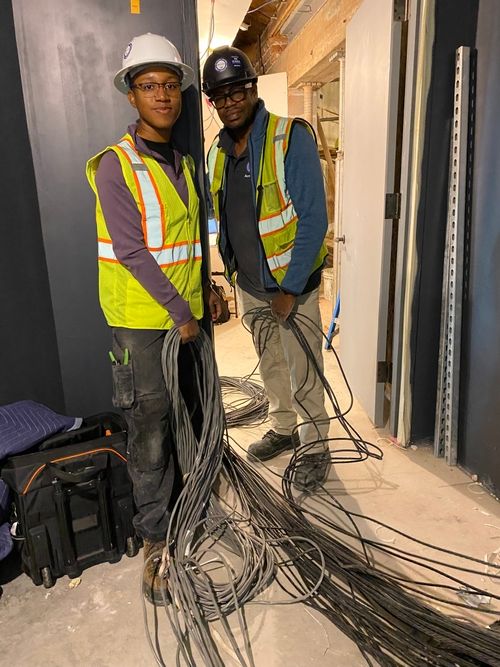 Two young black people wearing hardhats and high visibility vests are laying down a large bundle of thin cables.