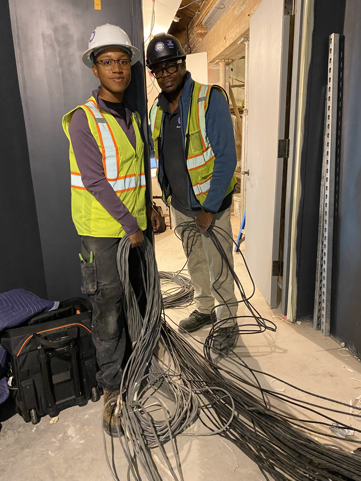 Two young black people wearing hardhats and high visibility vests are laying down a large bundle of thin cables. 