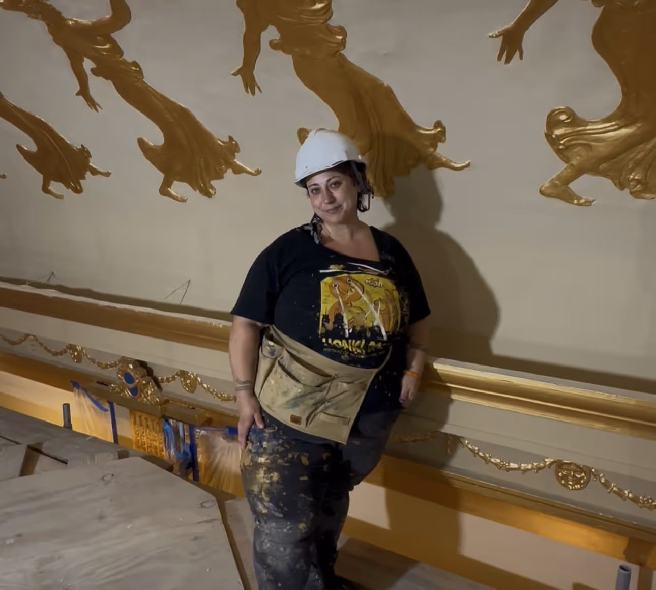 Helen McCarthy wearing jeans and a tee shirt, a white hard hat, and a large rectangular tool belt. She is pictured in front of the golden muses that line the Zeiterion ceiling. 