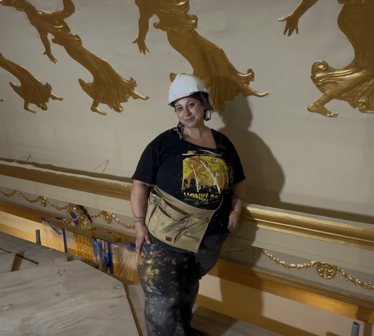 Helen McCarthy wearing jeans and a tee shirt, a white hard hat, and a large rectangular tool belt. She is pictured in front of the golden muses that line the Zeiterion ceiling. 