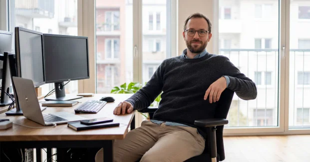 Professional AI Avatar presenter sitting at a desk with dual monitors in a bright office setting.