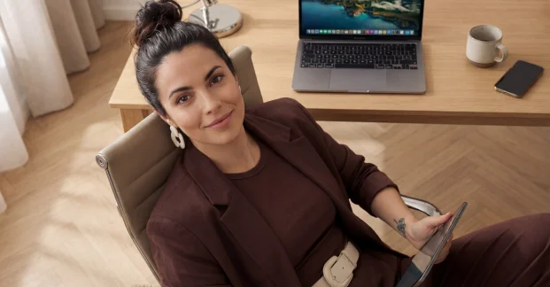Confident AI Avatar presenter seated at a desk holding a tablet in a warm office environment.