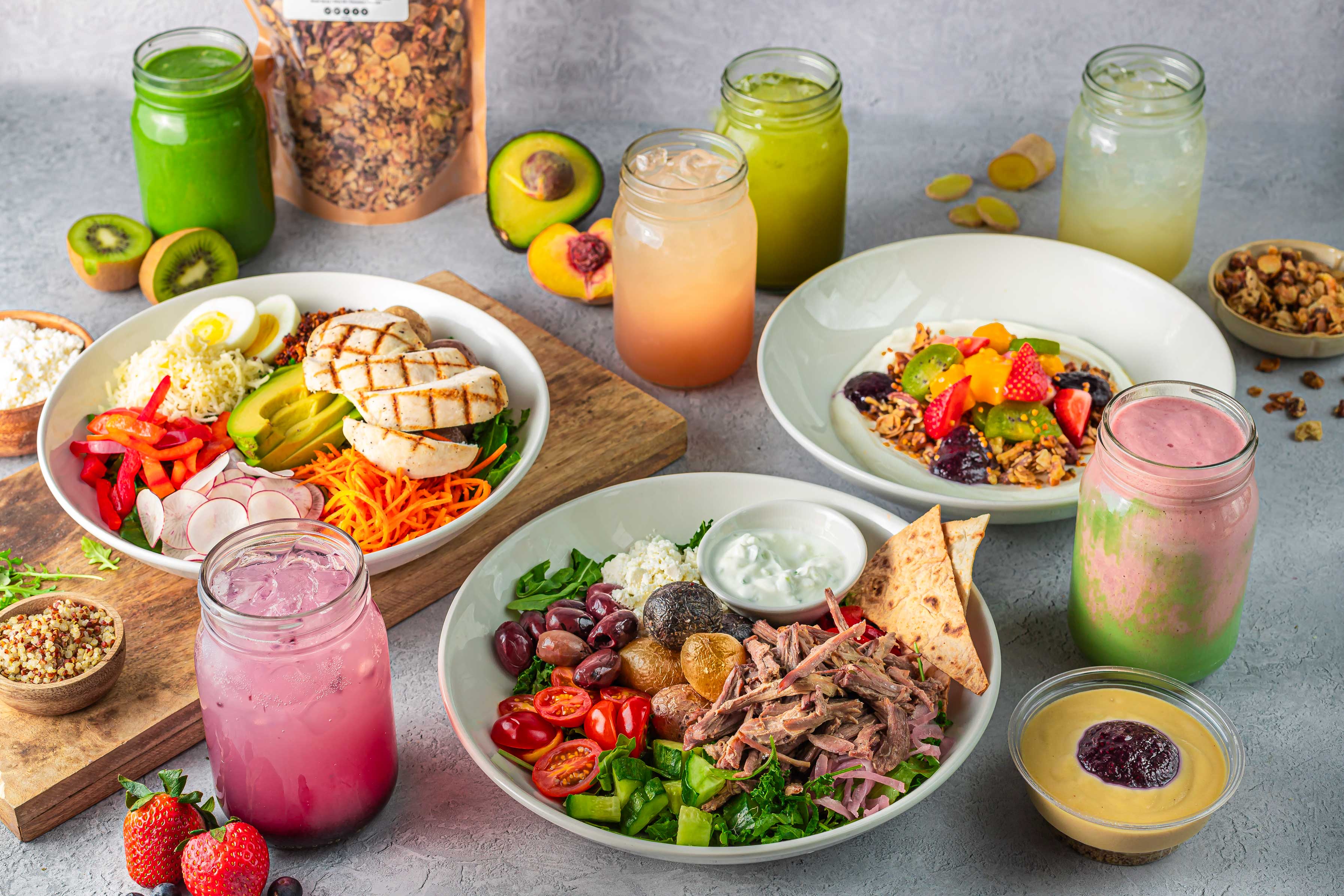 A variety of colorful healthy meals, including salads with grilled chicken and vegetables, fruit-topped yogurt bowls, smoothies in jars, green juice bottles, avocados, and granola on a gray tabletop.