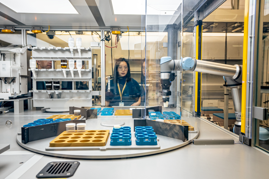 Dark-haired scientist in the center of the frame looks toward the camera. They are standing behind the clear glass of an encased automated lab.