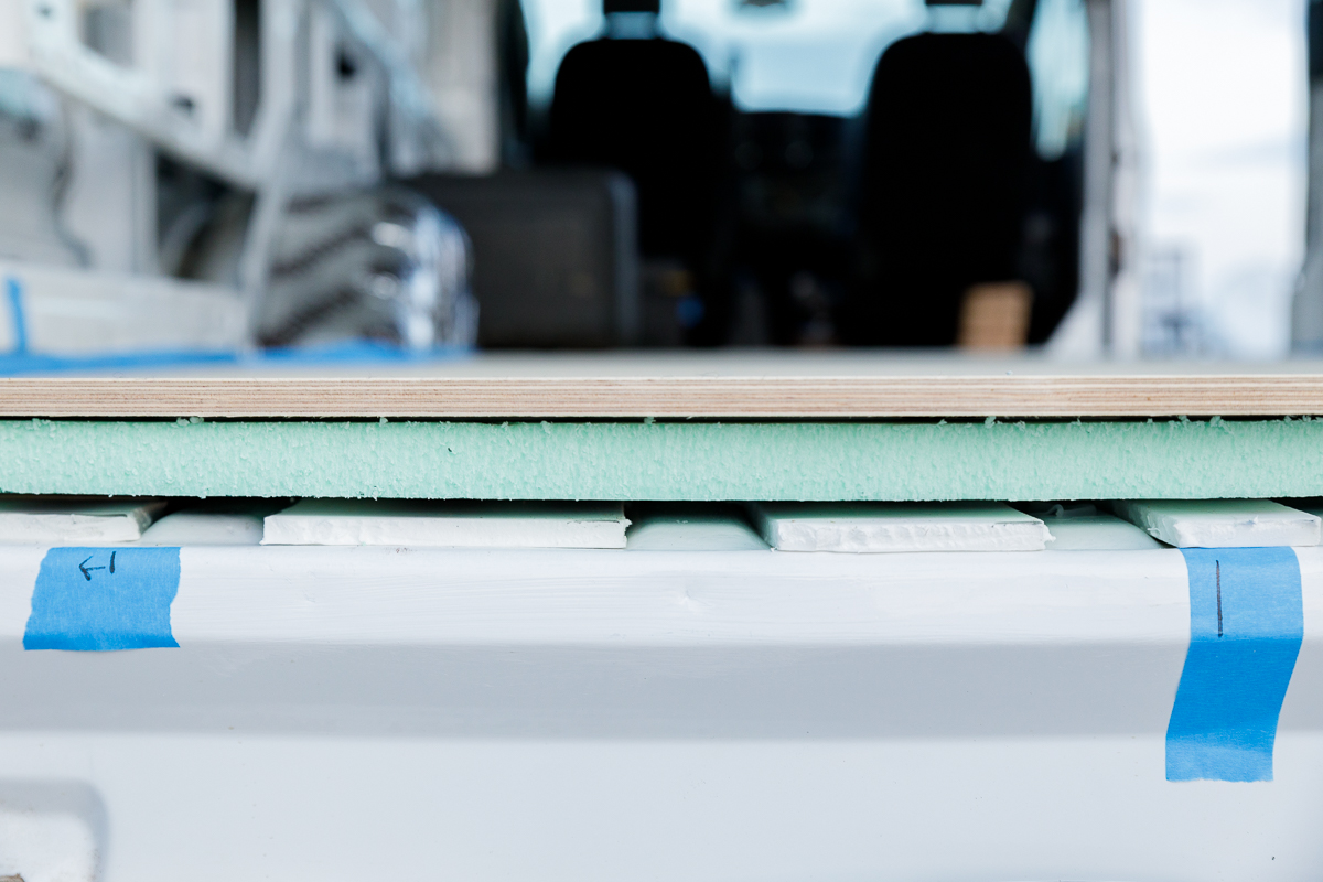 Close up on three layers of flooring in the rear of a campervan at eye level. One layer of white minicell foam is on the bottom, followed by a one inch thick green foam board layer and then half inch thick plywood