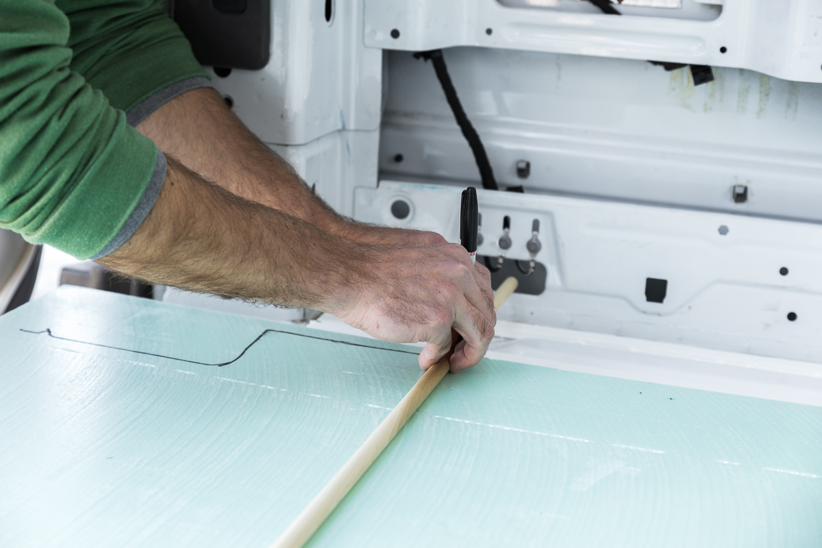 Close up on a man's hands as he uses a wood dowel rod and sharpie marker to scribe foam insulation board to the wall of a cargo van