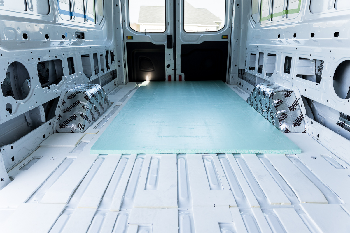 The floor of an empty cargo van with white minicell foam insulation filling the corrugations in the floor and a piece of green XPS foam board laying on top