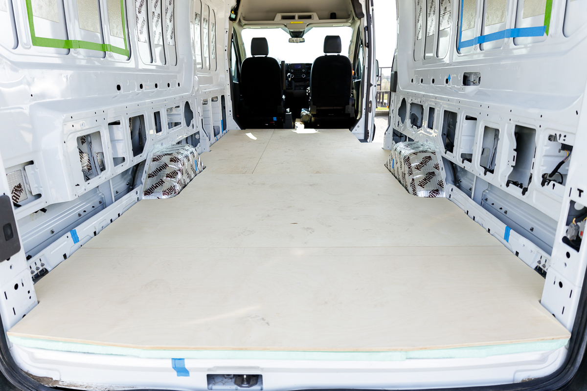 The rear of an empty cargo van being converted into a campervan with a plywood subfloor neatly set in place