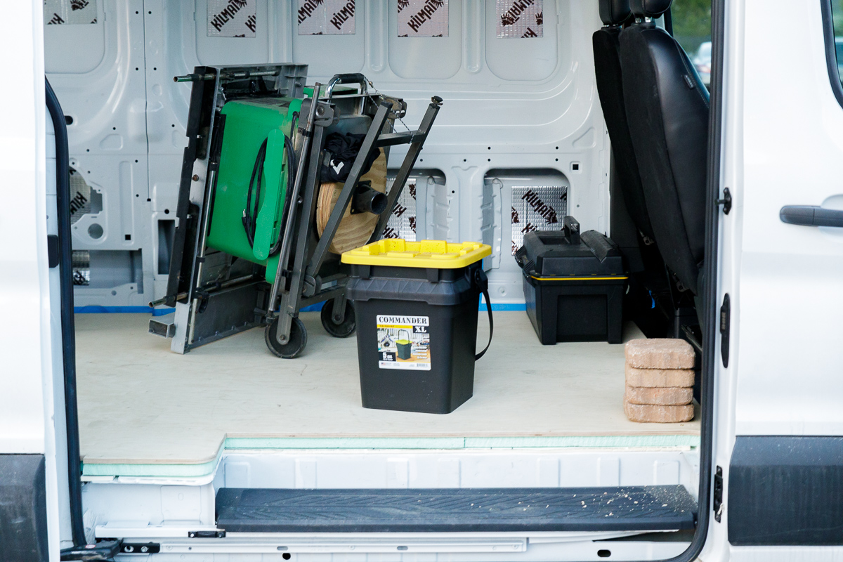 Heavy objects including bricks, a table saw, and containers being used to put weight on a plywood subfloor installed in the rear of a campervan conversion